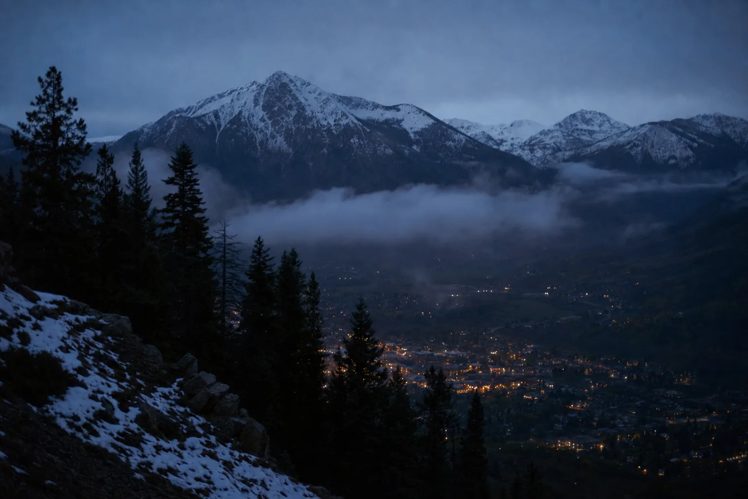 A Colorado mountain town at blue hour, fog settling in the valley below a ridge of pines